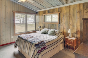 Un dormitorio con una cama grande, dos ventanas, techo de madera y una mesita de noche con lámpara.