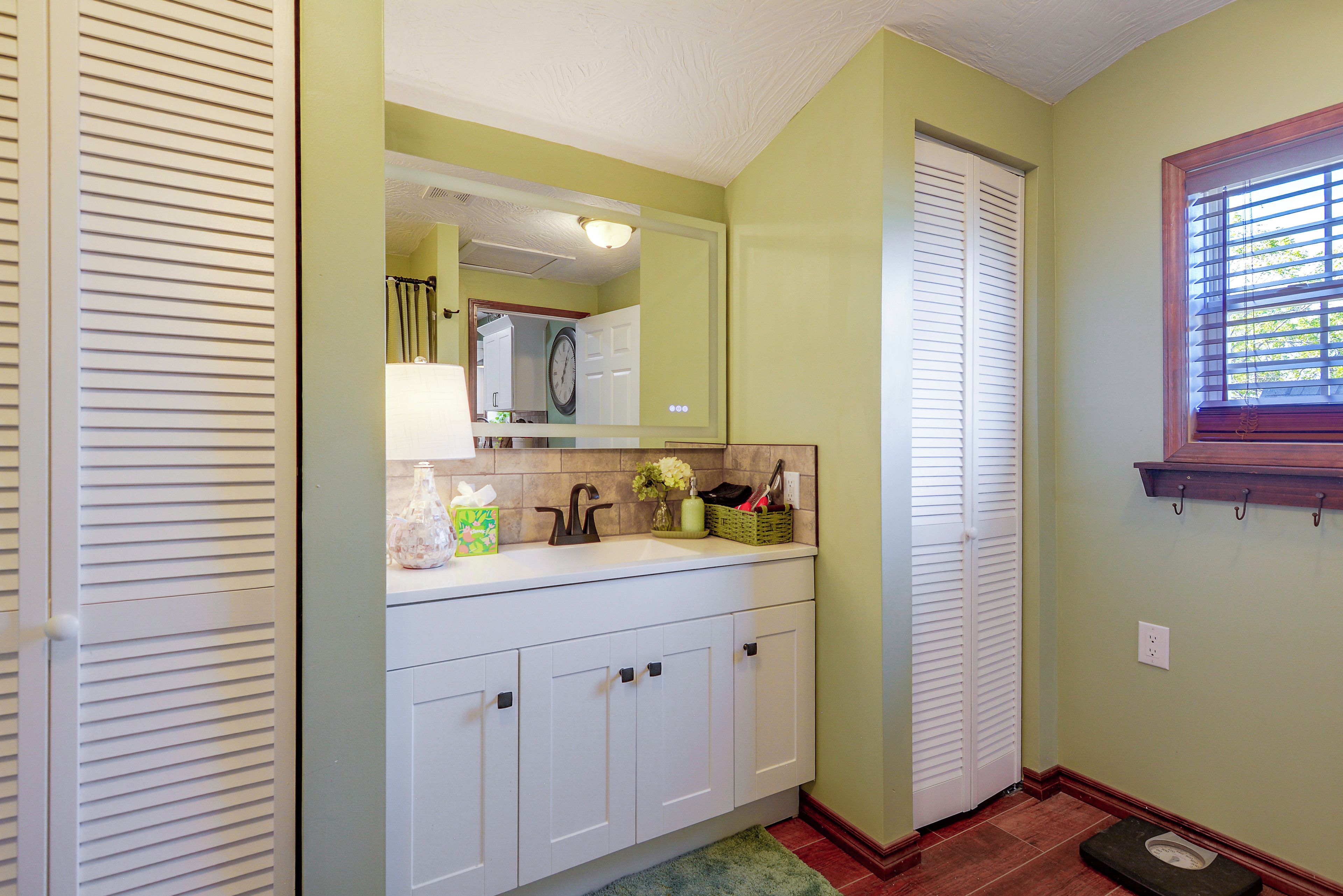 A bathroom with a white vanity, a mirror, a clock, and a window with blinds.