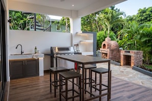 Cocina exterior con parrilla, taburetes de bar y horno de ladrillo, rodeada de abundante follaje.
