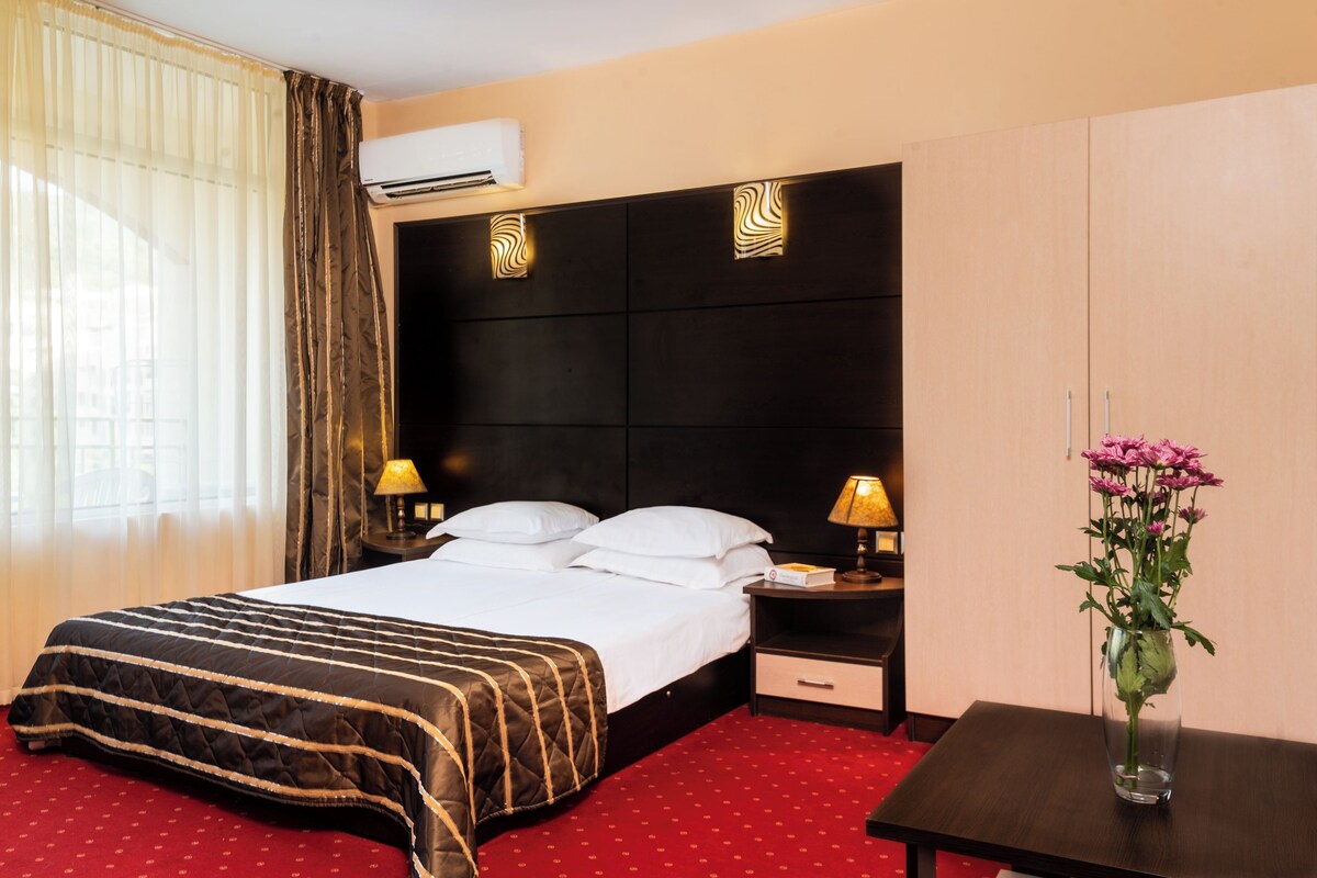A hotel room with a bed, two bedside tables, a nightstand, a vase with flowers, and an air conditioning unit.