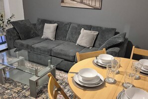 Ein modernes Wohnzimmer mit einem grauen, gestreiften Sofa, einem Glastisch und einem Holzespeisetisch, gedeckt mit Tellern und Gläsern.