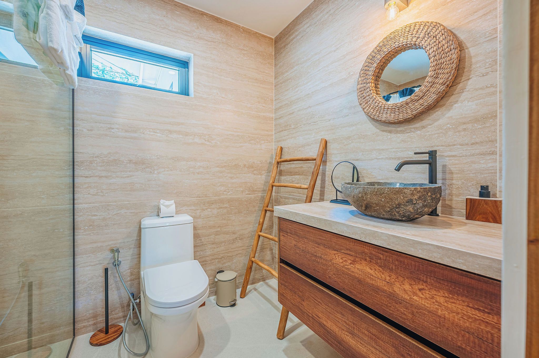 Une salle de bain avec des toilettes, une échelle en bois, un miroir rond et un lavabo en pierre.