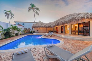 A swimming pool with lounge chairs, a thatched-roof structure, and a dining area with a view of the pool. - Your private Aruba escape with nearly everything brand new, 4 bedrooms, a tropical pool, waterfall wish spot, BBQ, and easy access to Palm Beach. (Noord)