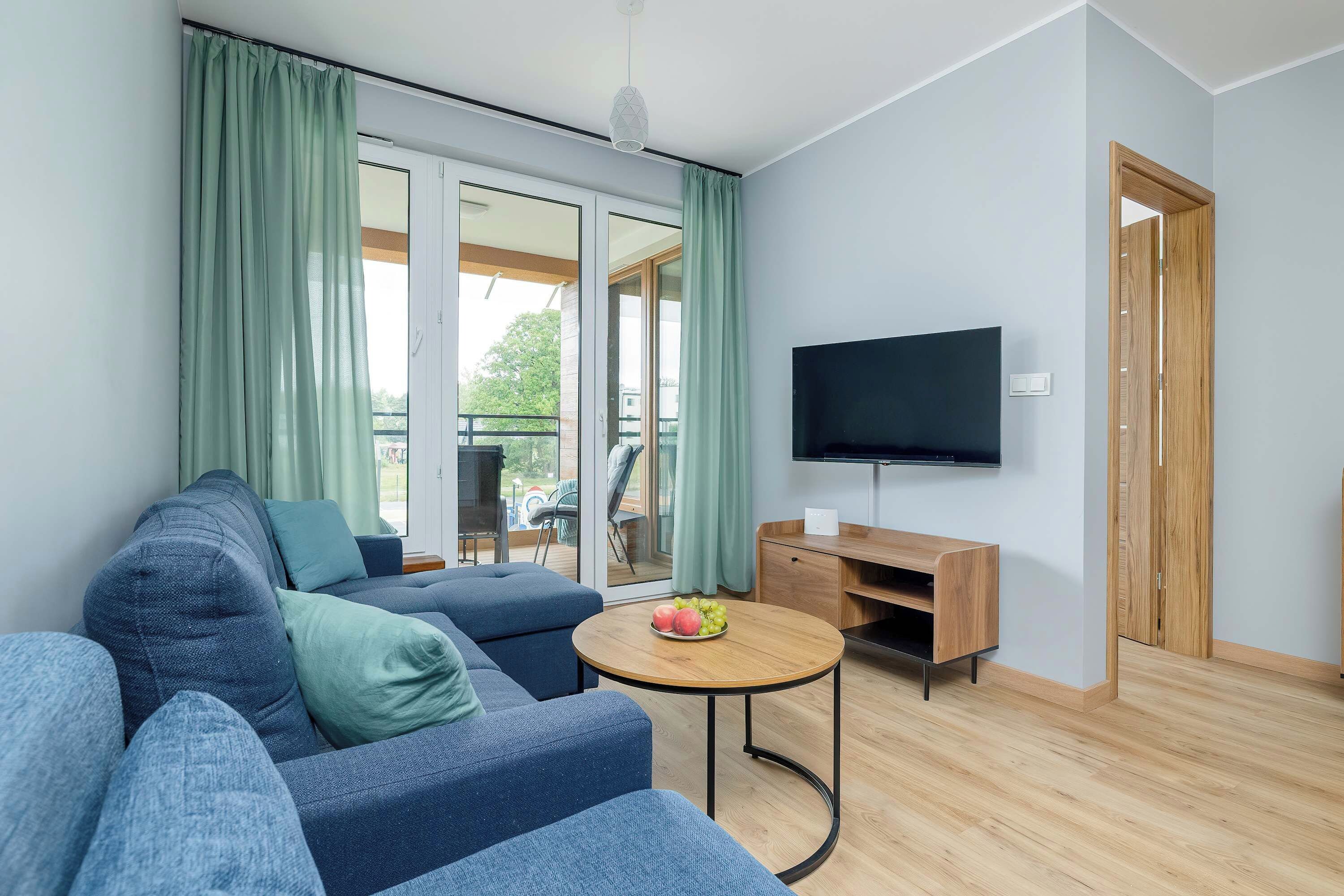 A modern living room with a sectional sofa, a coffee table with fruit, a flat-screen TV, and a sliding door leading to an outdoor area.