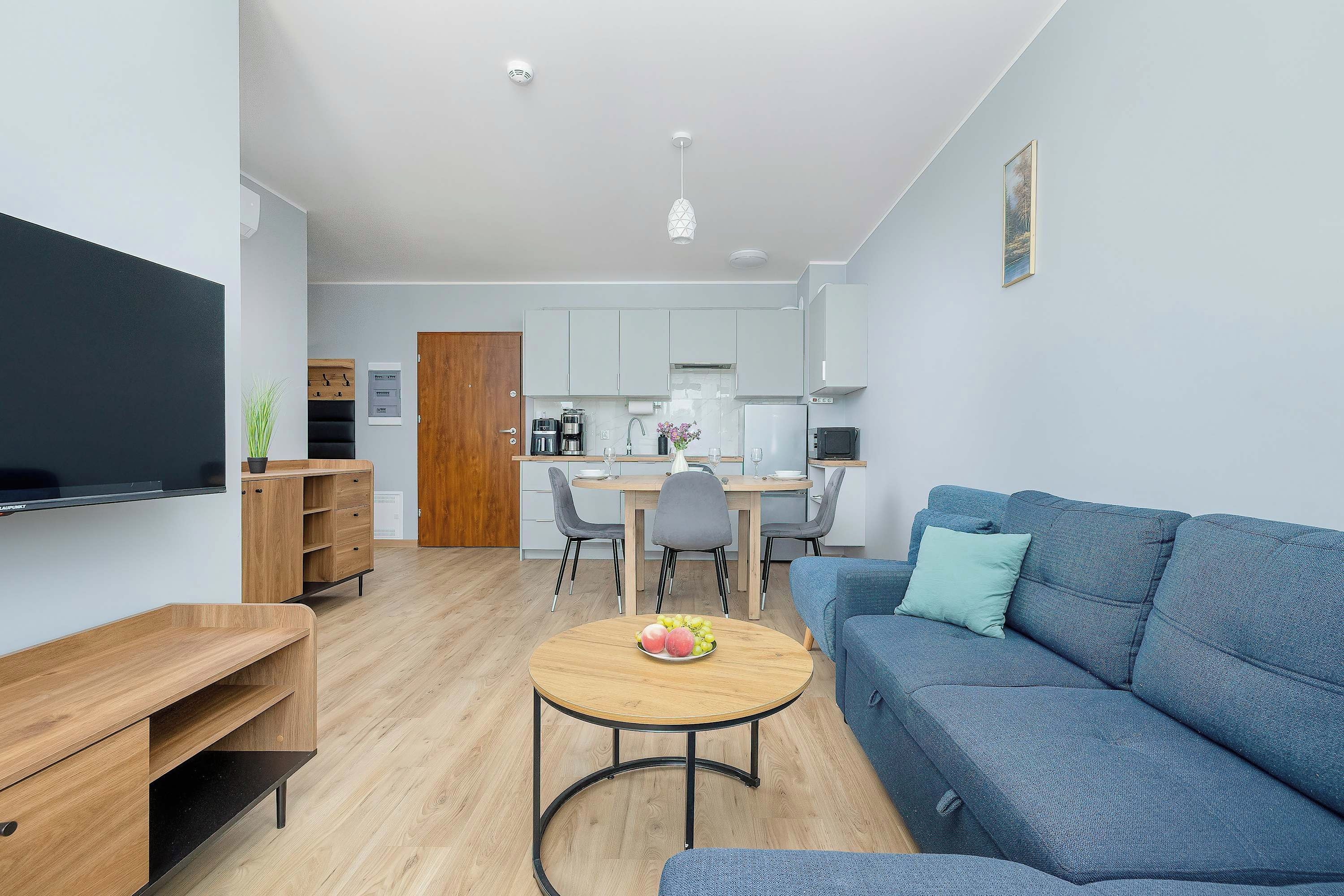 A modern living room with a blue sofa, a wooden coffee table with fruit, a TV, and a kitchen area with white cabinets and a dining table.