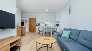 A modern living room with a blue sofa, a wooden coffee table with fruit, a TV, and a kitchen area with white cabinets and a dining table.