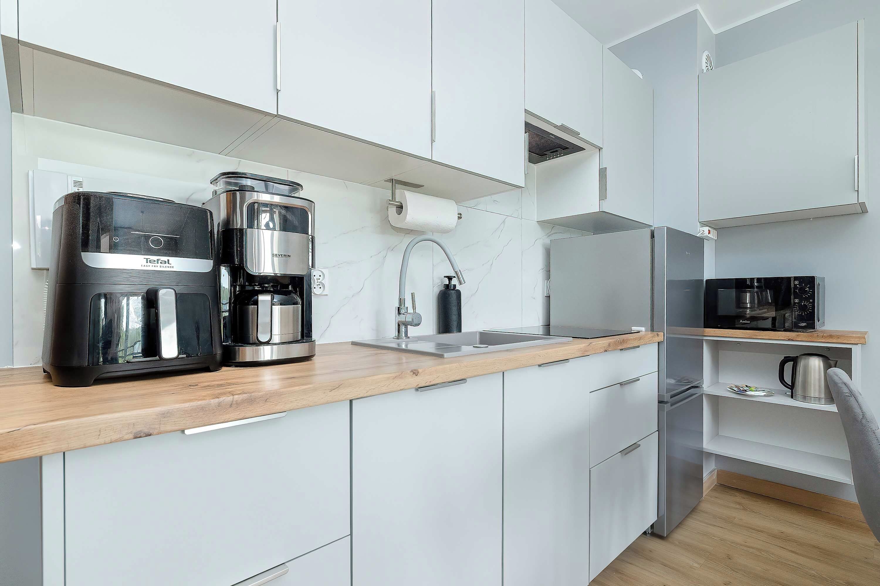 A modern kitchen with white cabinetry, a wooden countertop, a coffee maker, a kettle, and a microwave.