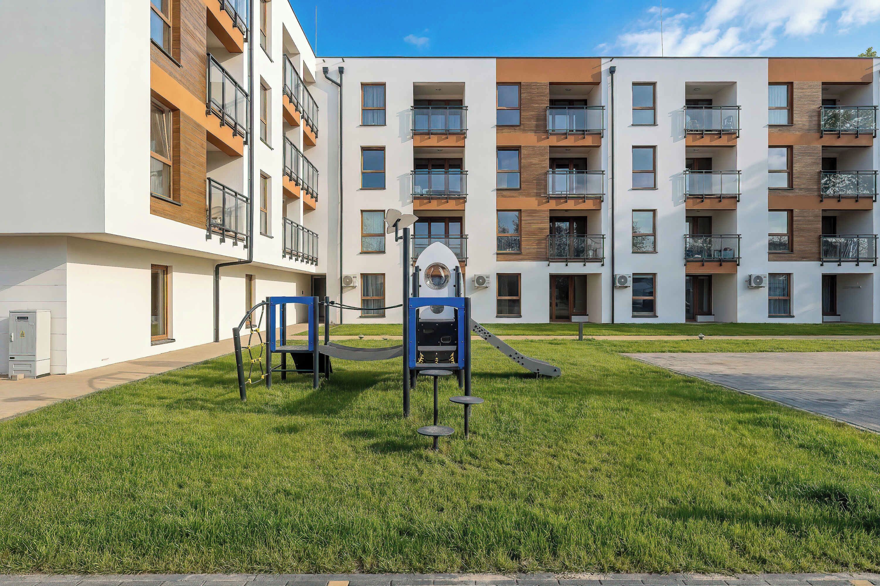 Een speeltuin met een glijbaan en klimconstructies, omgeven door een verzorgd gazon, voor een meerlaags woongebouw met balkons.