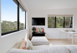 A modern living room with a large window, a flat-screen TV, a desk, and a seating area with cushions. - The Lookout – Coastal Studio for Two (Waiheke Island)