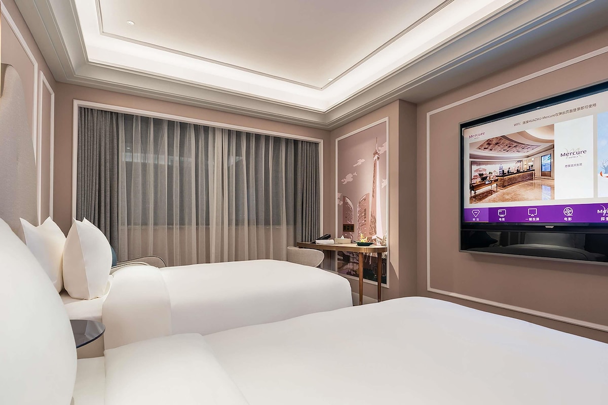Habitación de hotel con una cama grande, televisión montada en la pared, una mesita pequeña y ventana con cortinas.