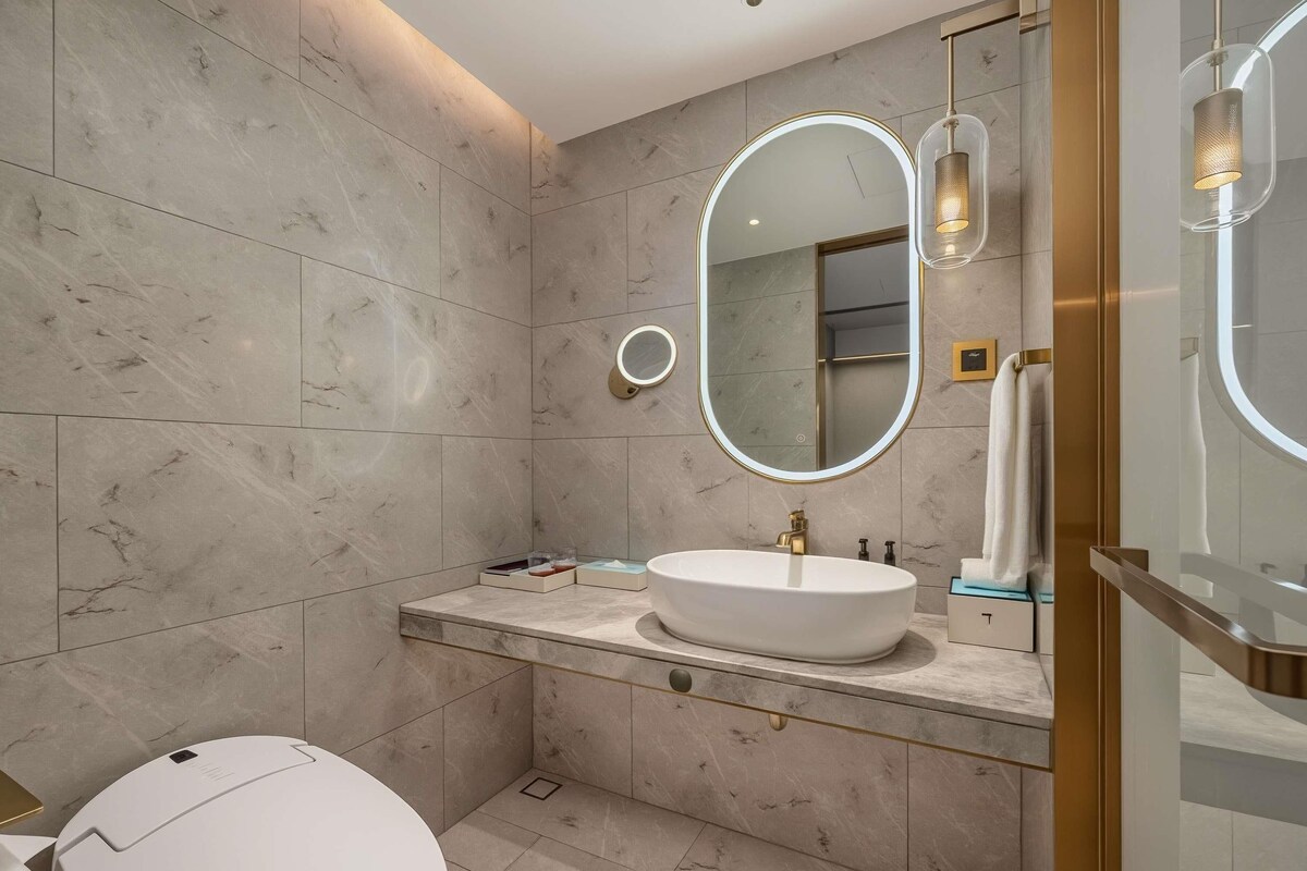 Un baño moderno con un inodoro blanco, un espejo ovalado grande y un lavamanos blanco con grifo dorado.