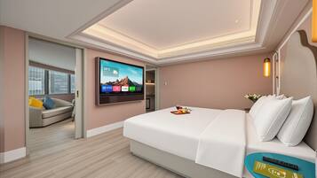 Habitación de hotel moderna con una cama grande, un televisor de pantalla plana montado en la pared, una mesita de noche con una bandeja de frutas y una copa de vino, y vistas a la ciudad a través de la ventana.