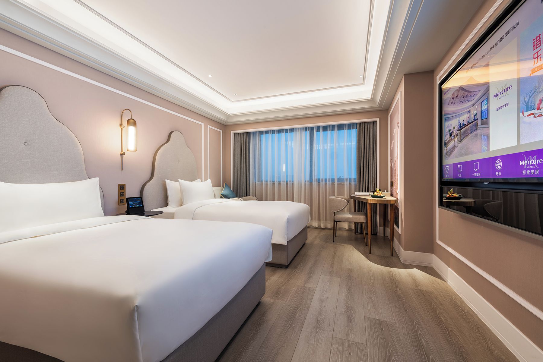 A hotel room with two beds, a desk, a television, and a view of the city.