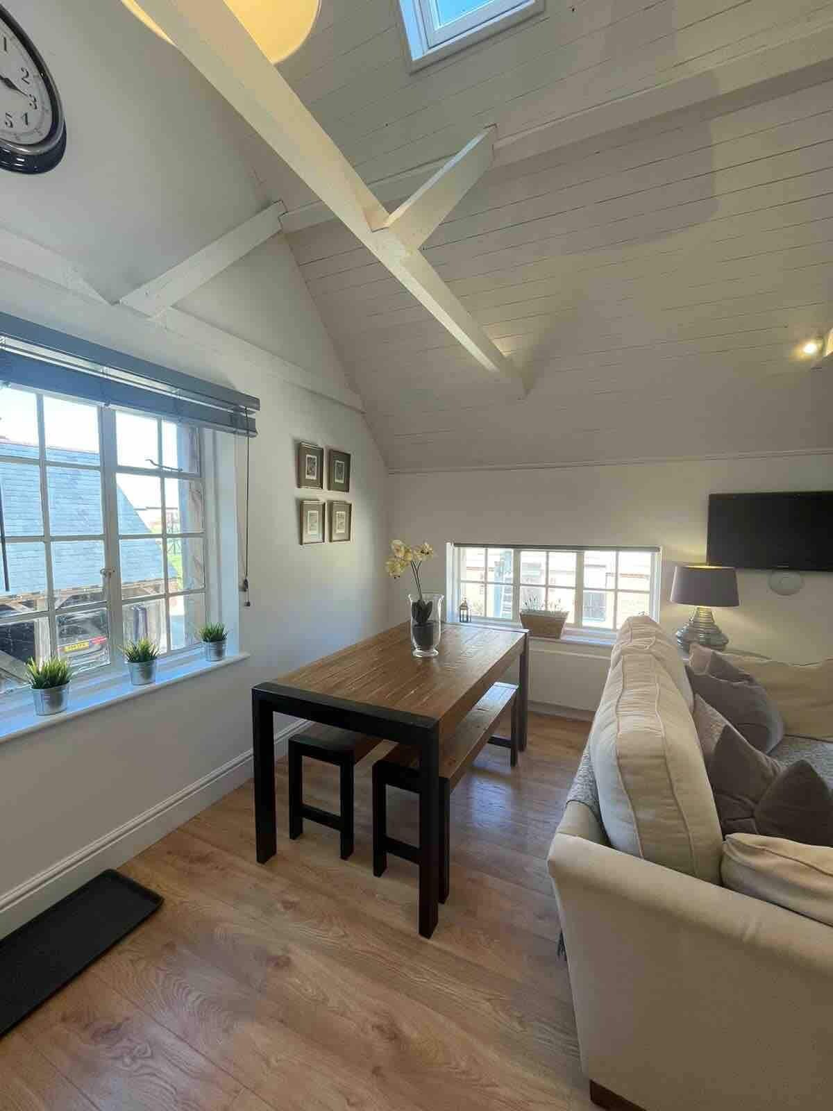 Een ruime loft met hoge plafonds, een houten eettafel, een bank en uitzicht op zee.