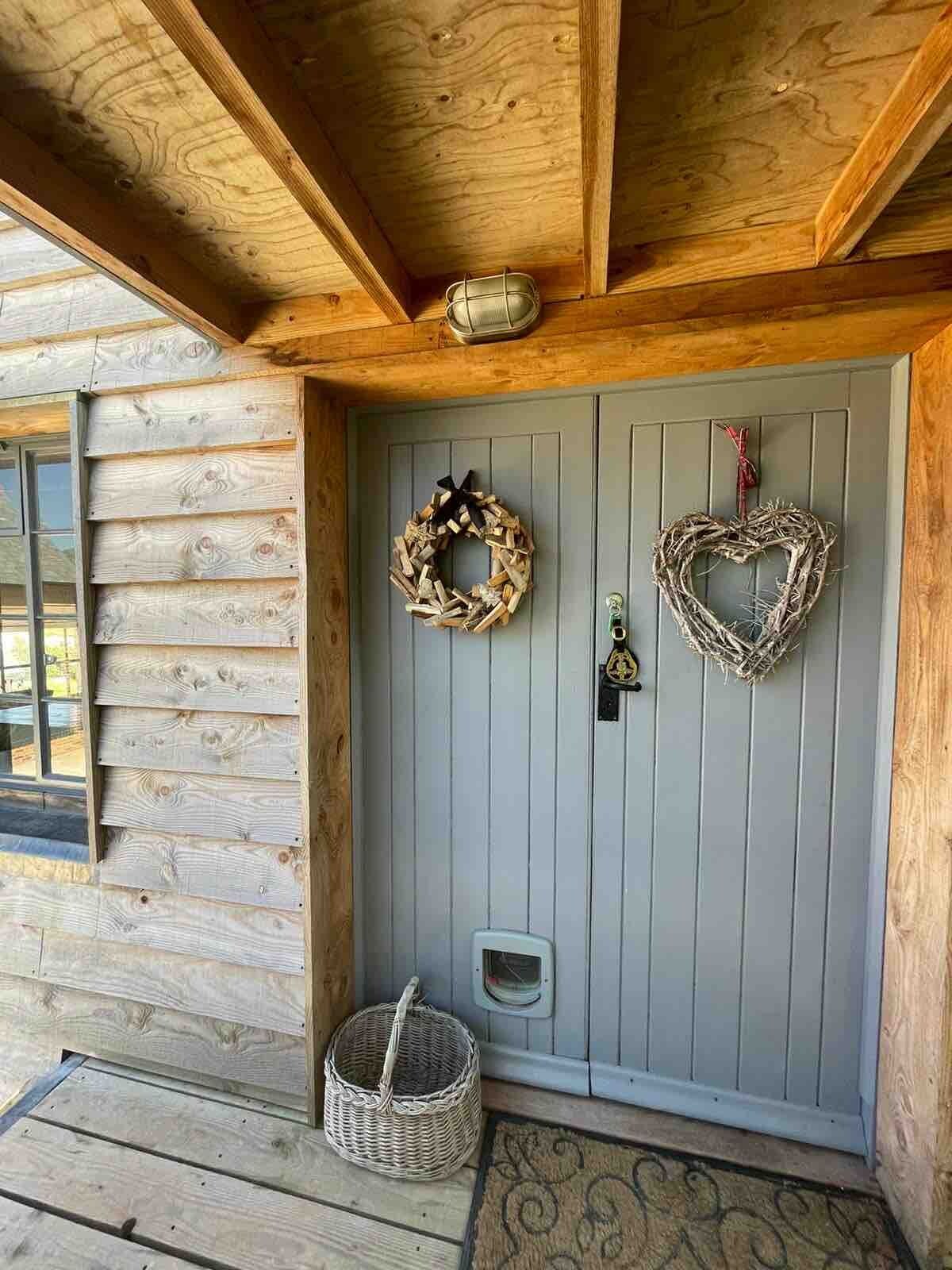 Entrada de una cabaña de madera con una puerta gris, dos coronas decorativas, una cesta y un ventanal pequeño.