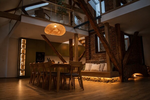 A spacious loft with exposed wooden beams, a dining area with a round table and chairs, and a cozy seating area with cushions.