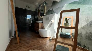A modern bathroom with a wooden floor, a white toilet, a sink, and a mirror. There is a window with a view of trees and a mountain.