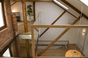 A loft-style room with exposed wooden beams, a glass railing, a staircase, and a hanging light fixture.