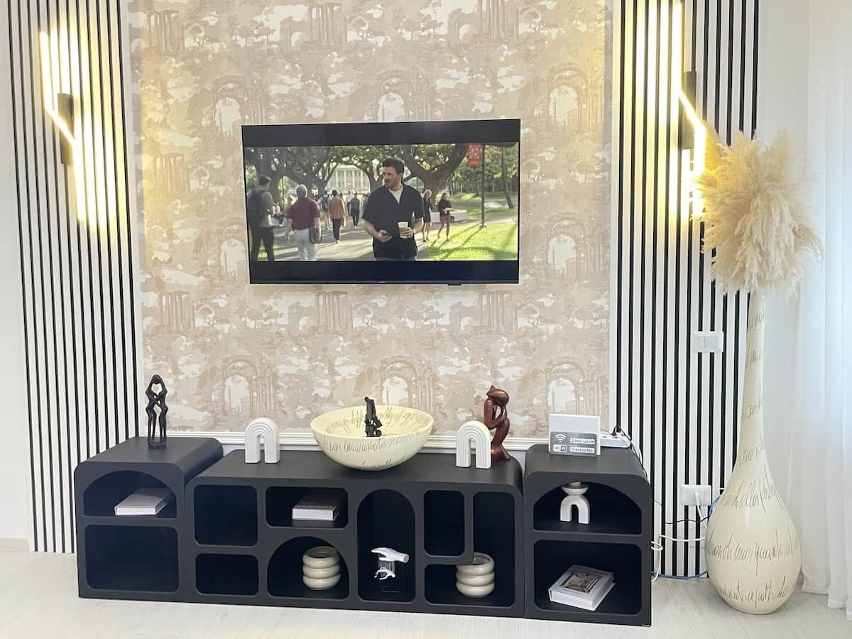 Een moderne woonkamer met een flatscreen televisie aan de muur, een zwarte entertainmentkast met planken, een decoratieve vaas en behang met een patroon.