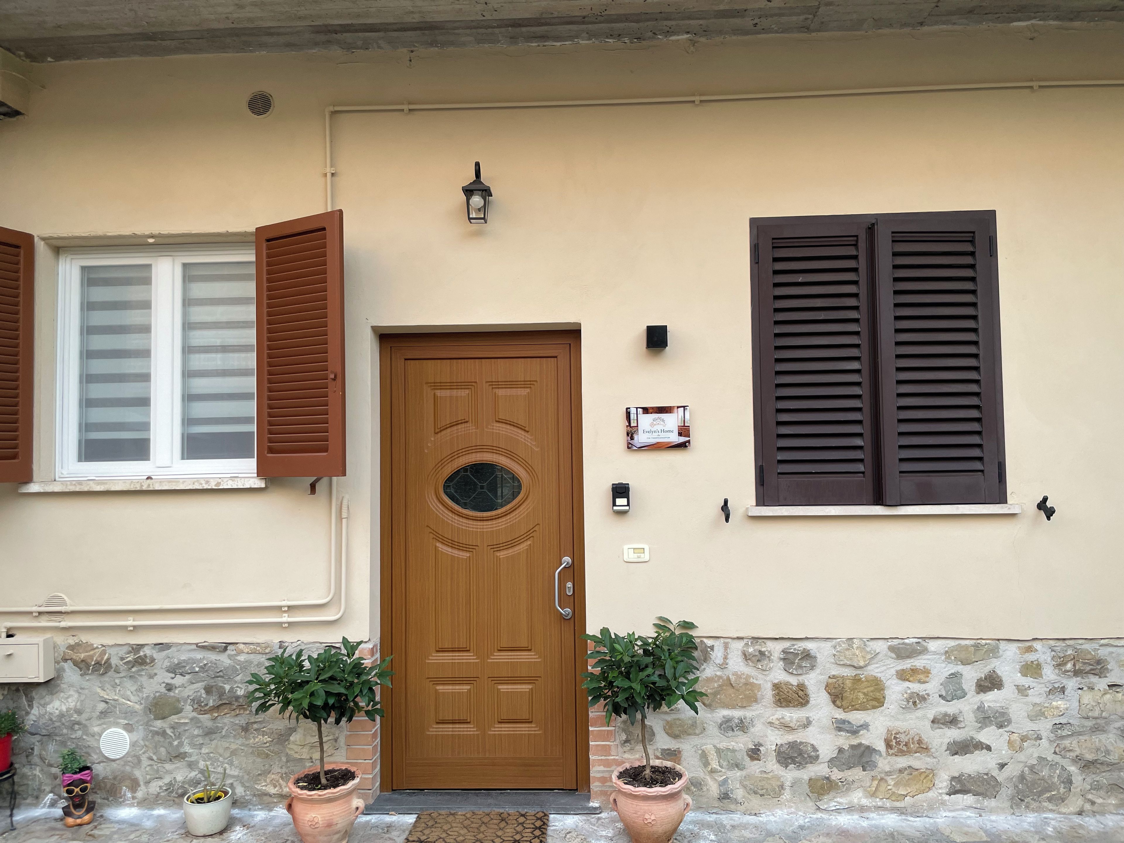 Un edificio color crema con puerta de madera, contraventanas marrones y dos macetas.
