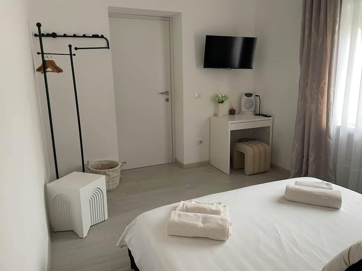 A hotel room with a bed, a TV mounted on the wall, a desk with a chair, a small table with a plant, and a coat rack with clothes.