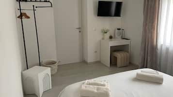A hotel room with a bed, a TV mounted on the wall, a desk with a chair, a small table with a plant, and a coat rack with clothes.