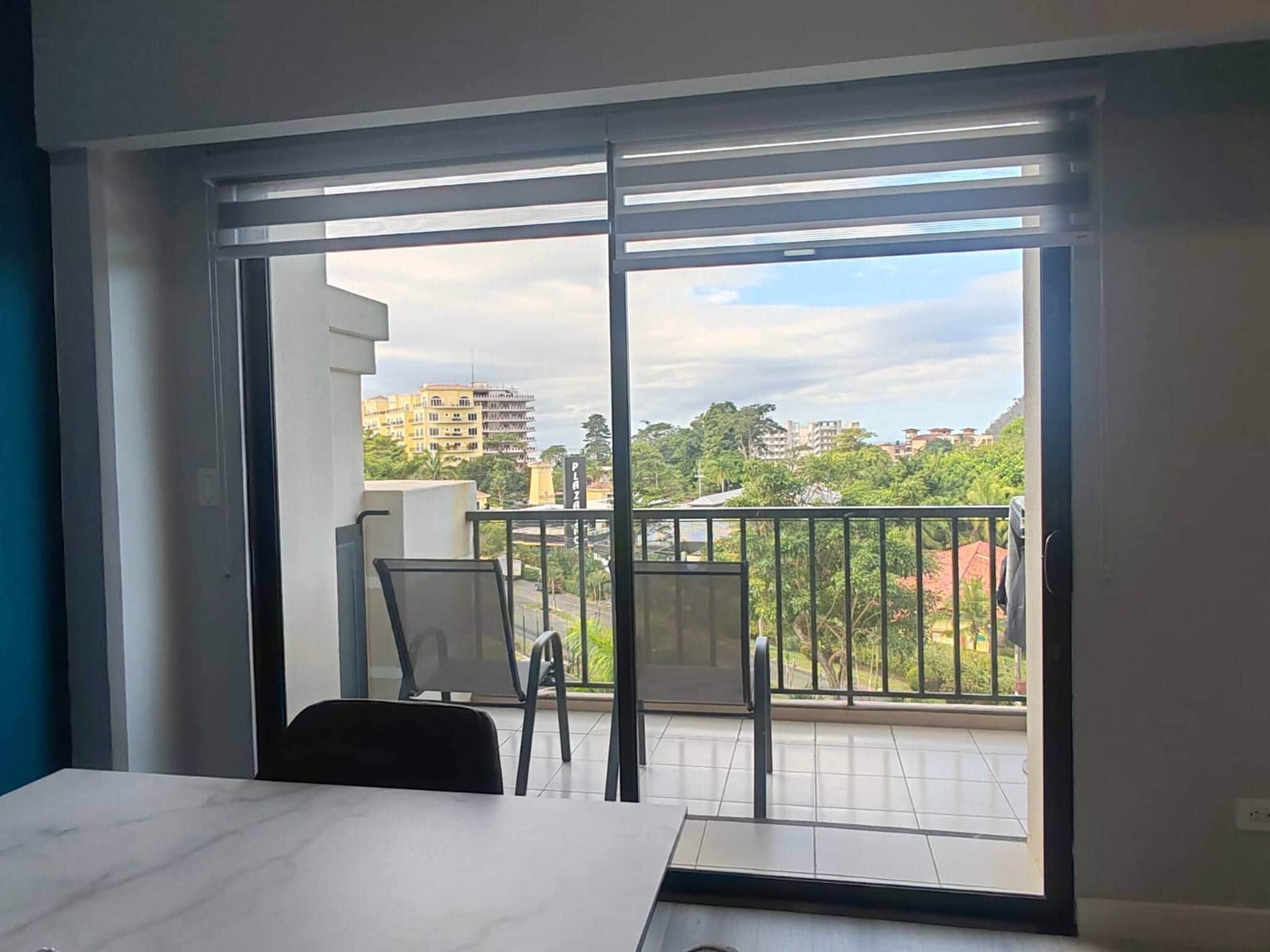 Ein Zimmer mit einer großen Schiebeglastür, die auf einen Balkon mit Blick auf Gebäude und Bäume führt.
