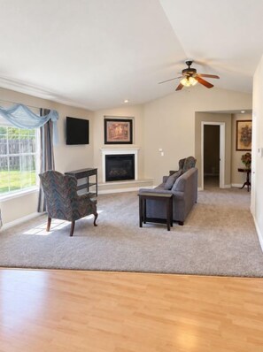 A spacious living room with a fireplace, a ceiling fan, and a large window.