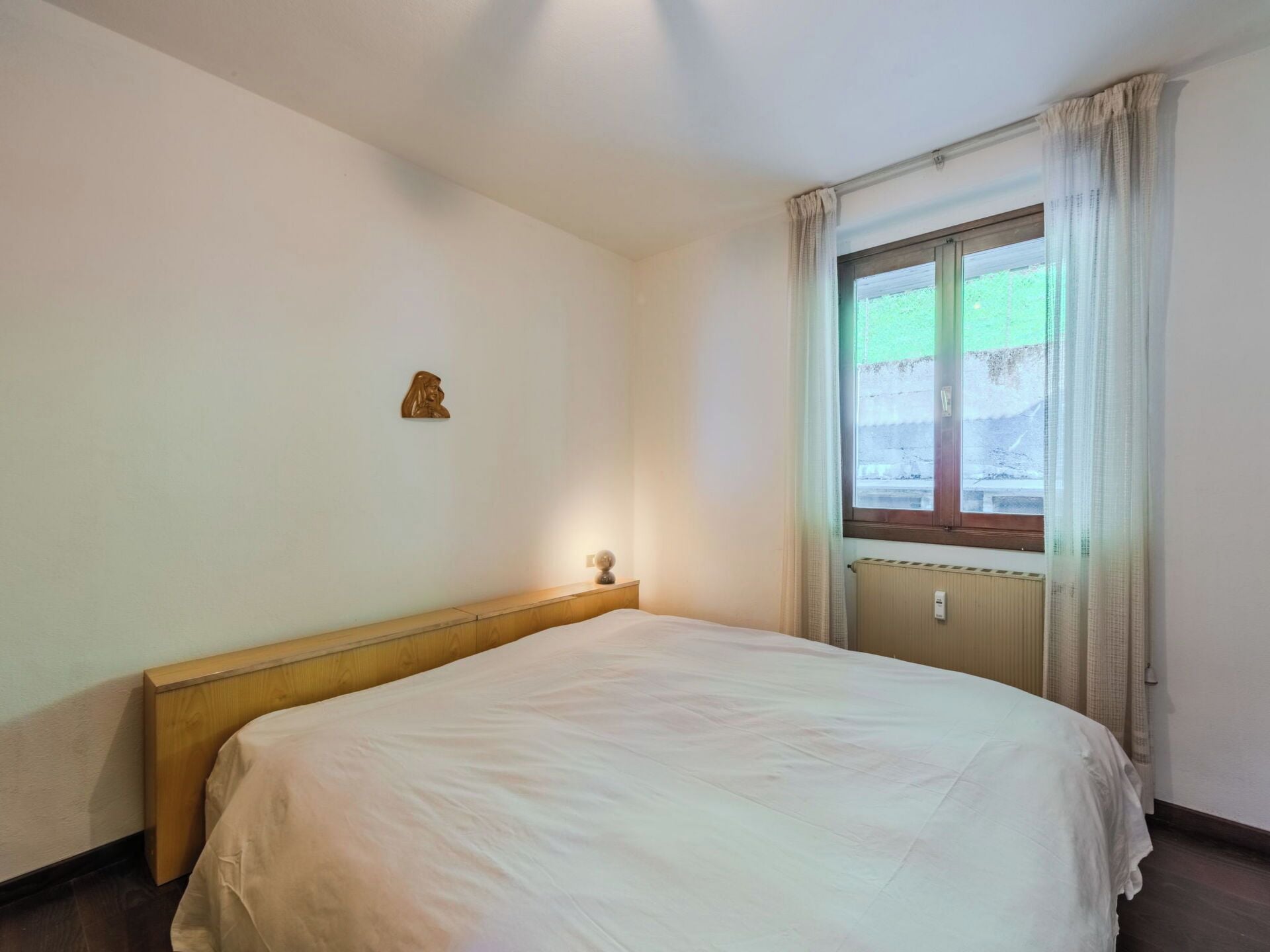 Een eenvoudige kamer met een enkel bed, een nachtkastje, een wandlamp en een raam met gordijnen.