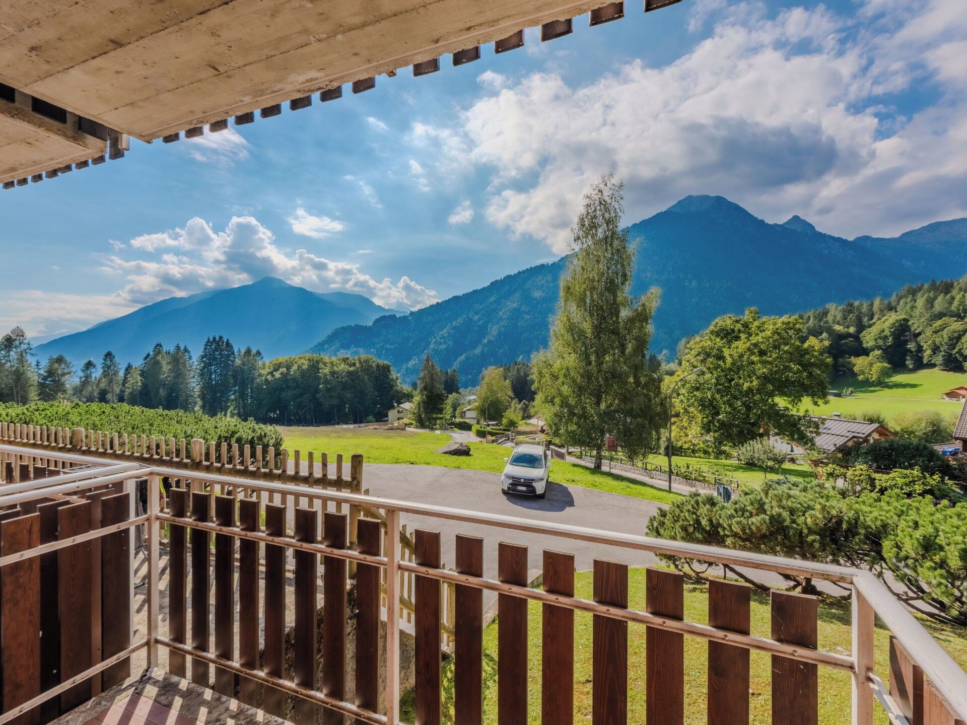 A balcony with a view of a mountain landscape, a road with a car, and a wooden fence.