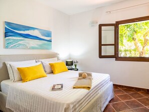 A neatly made bed with white bedding and two yellow pillows, a painting of a wave on the wall, and a window with a view of trees. - Wonderful private villa for 4 people with A/C and TV (San Teodoro)