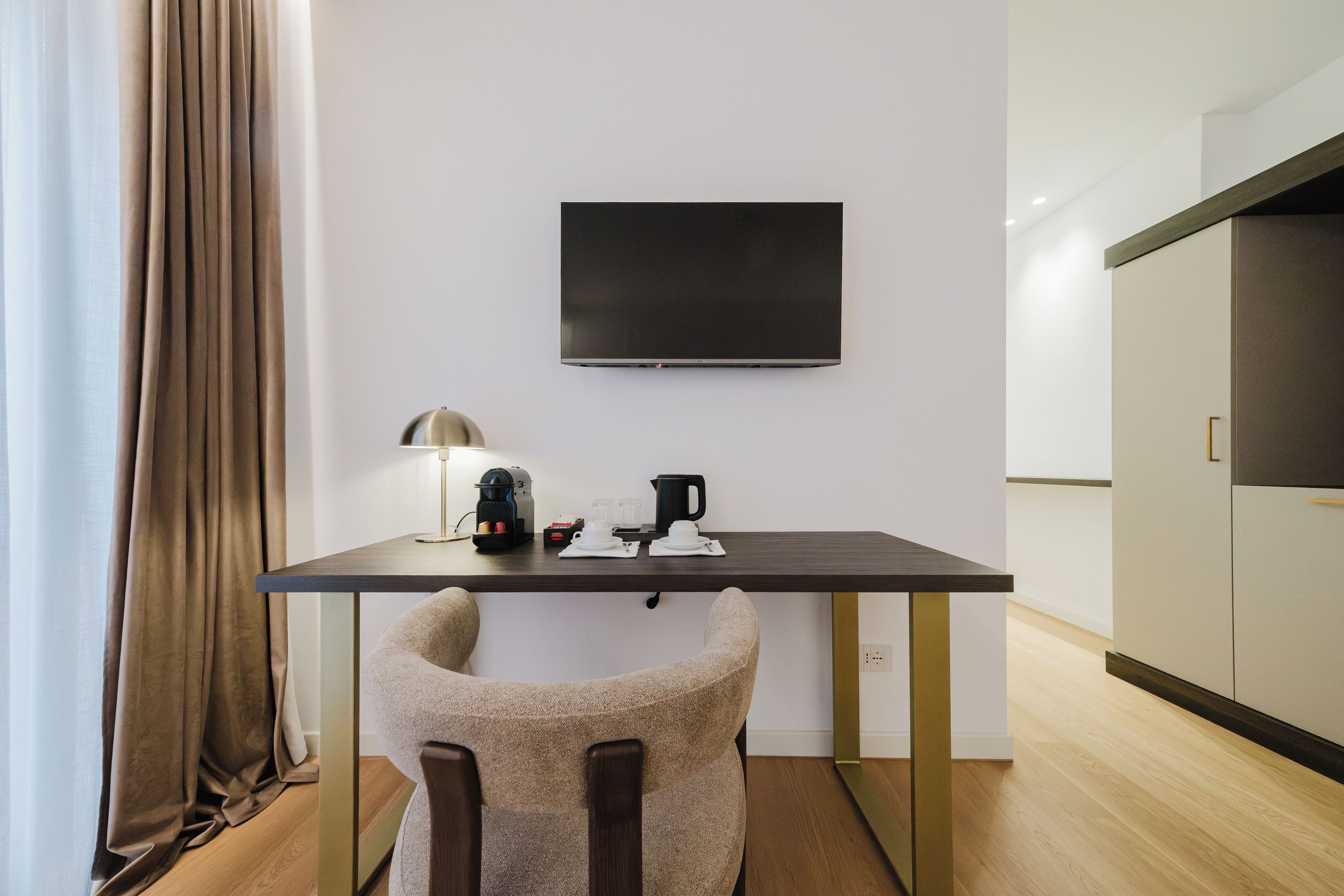 A small, modern room with a flat-screen TV mounted on the wall, a table with a coffee maker, cups, and a lamp, and a chair with a unique design.