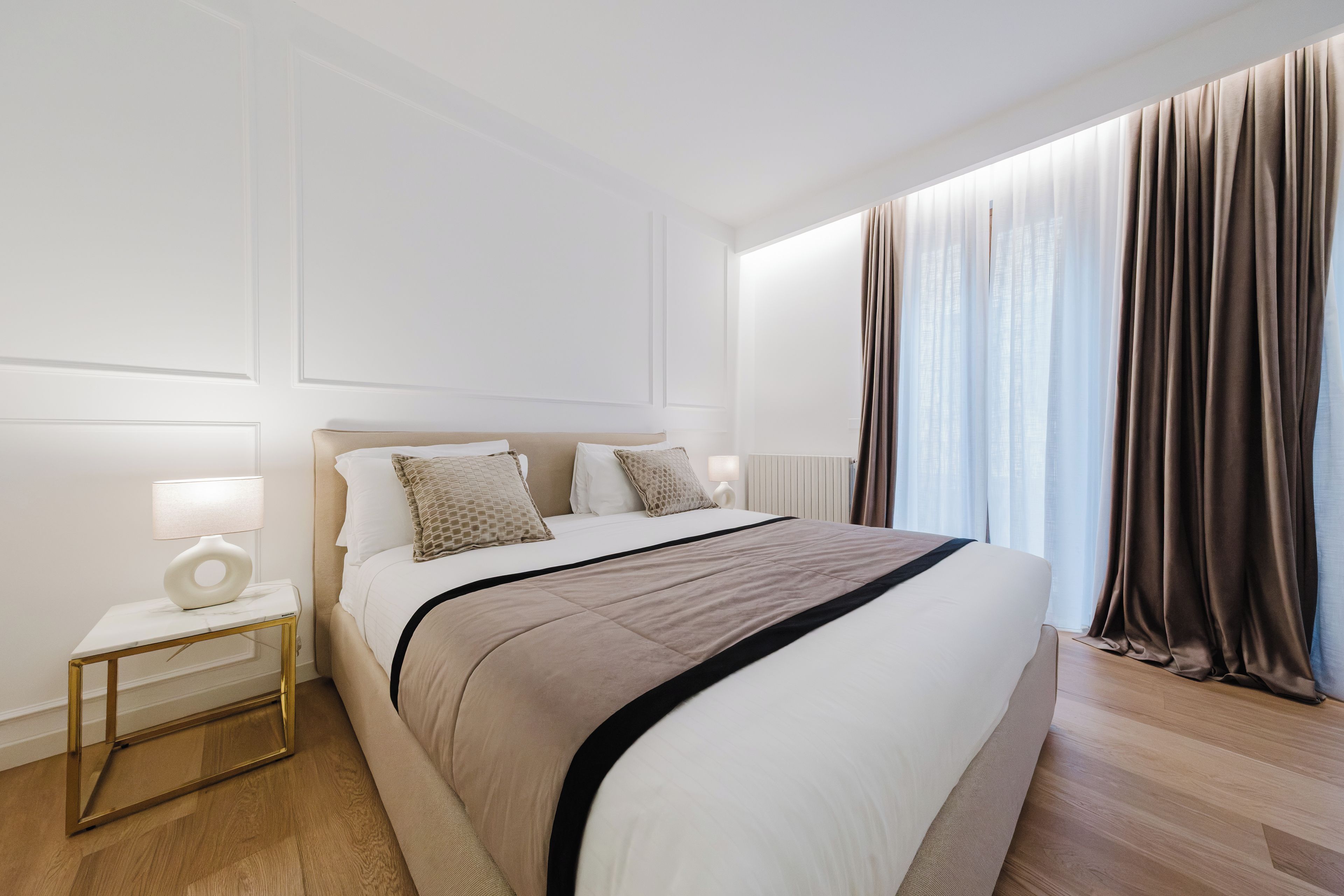 Een moderne hotelkamer met een groot bed, nachtkastjes en uitzicht op de stad door dunne gordijnen.