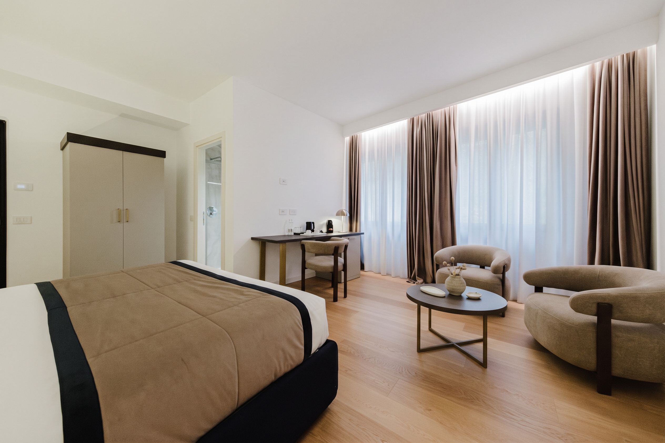 A hotel room with a bed, a desk, two armchairs, and a large window with curtains.