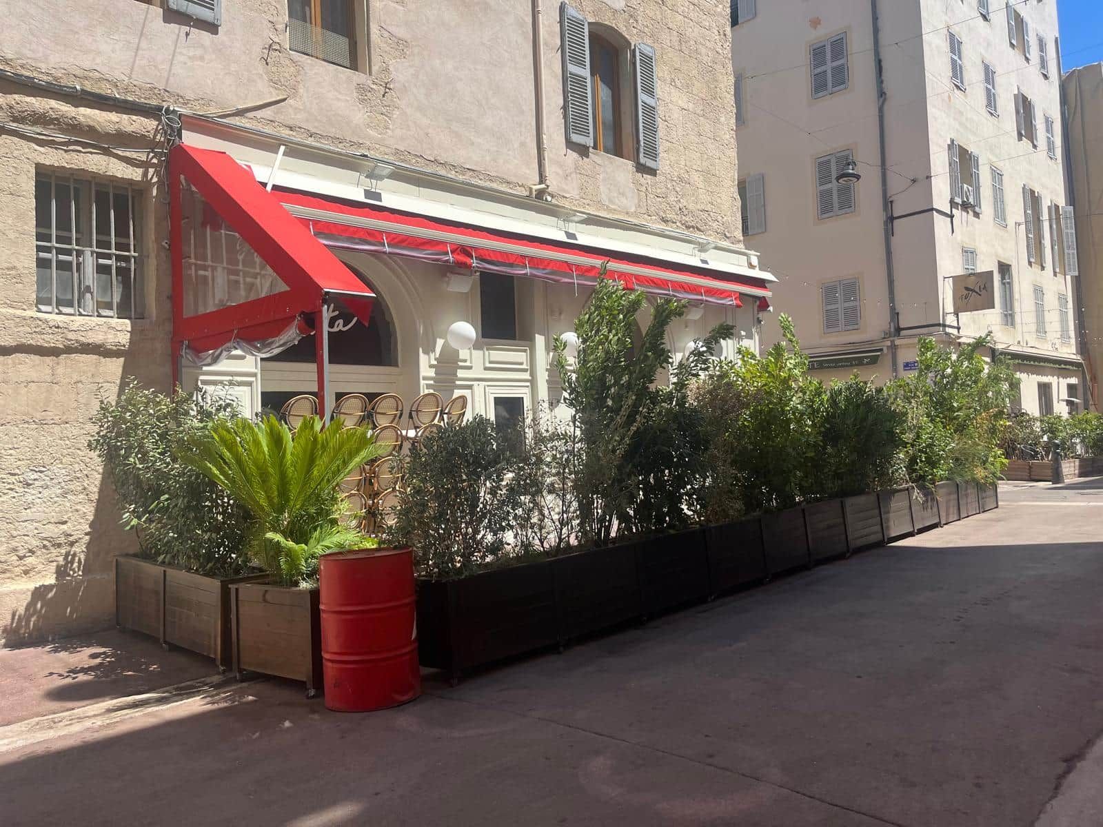 Un edificio con un tendone rosso, posti a sedere all'aperto con sedie in vimini e una botte rossa.