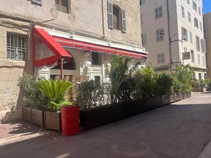 Un edificio con un tendone rosso, posti a sedere all'aperto con sedie in vimini e una botte rossa.