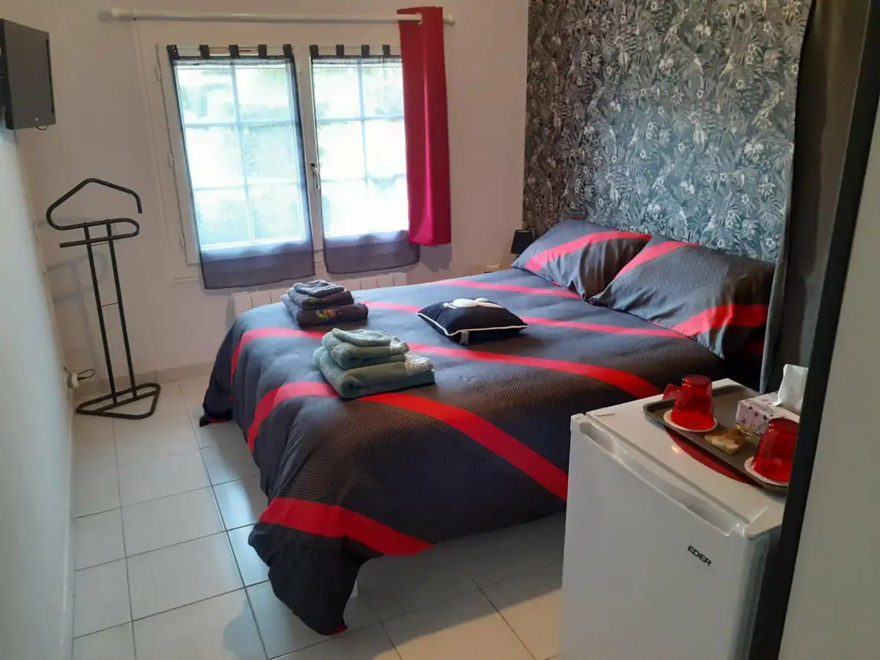 A bedroom with a bed, a small fridge, a window with curtains, and a wall with a floral pattern.
