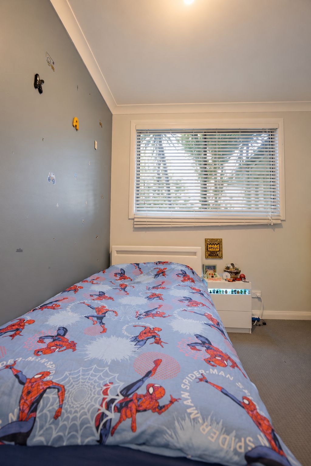 A bedroom with a bed featuring a Spider-Man themed bedspread, a window with blinds, a radiator, and a small shelf with items.