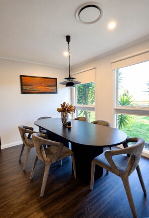 A dining area with a round table, six chairs, a pendant light, a framed painting, and a window with a view of greenery. - Stylish Family Retreat with Sauna, Gym, Basketball & Alfresco (Saint Clair)