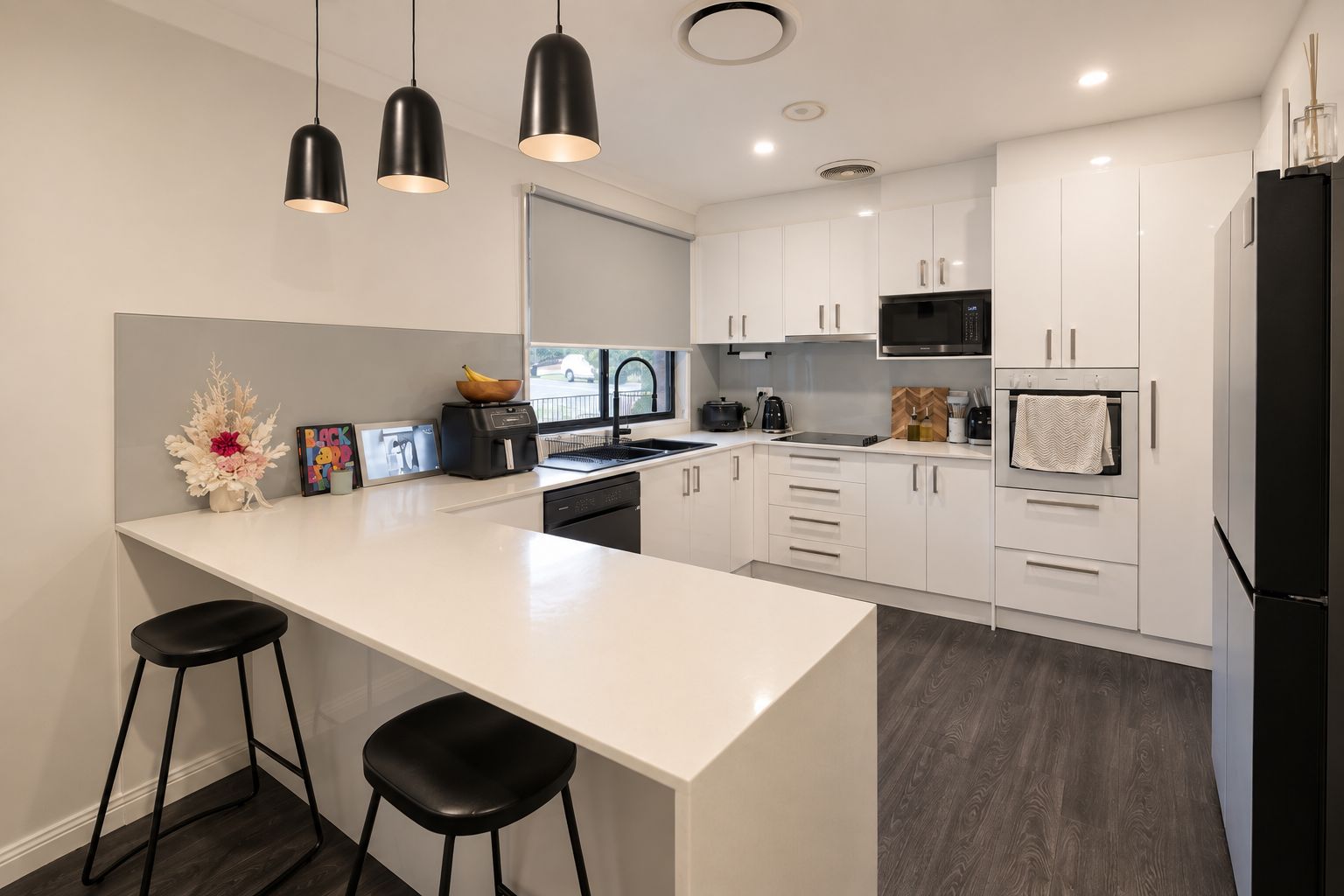 Cocina moderna con armarios blancos, una isla central con taburetes, iluminación colgante negra y vistas a la calle.