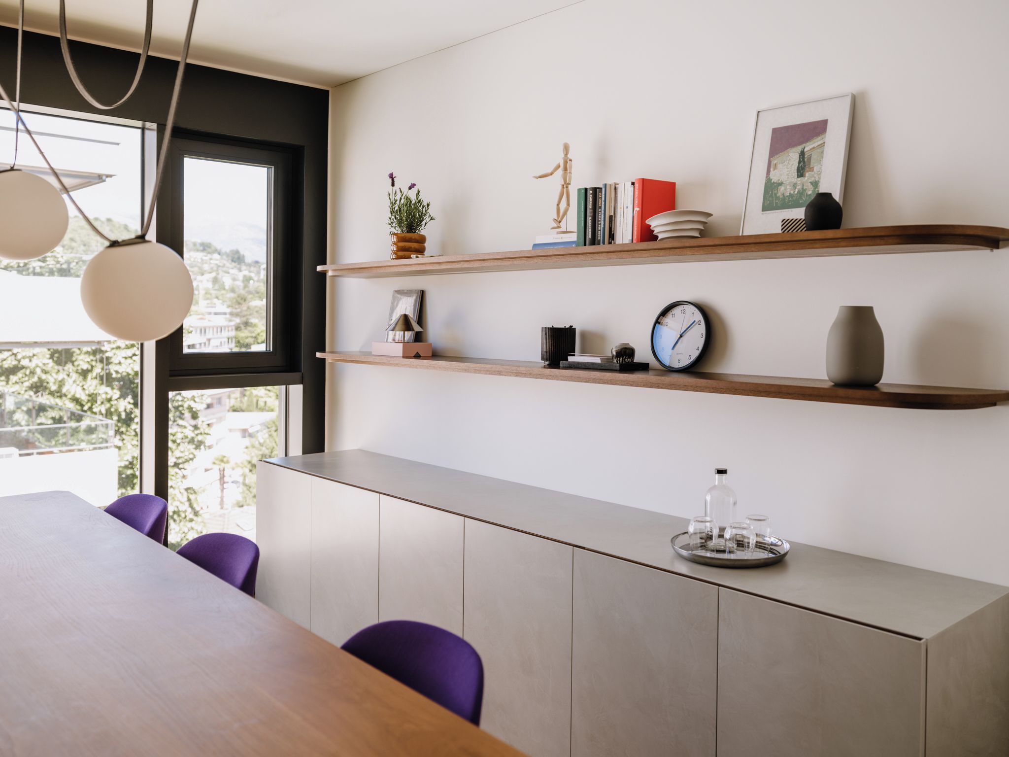 Un comedor moderno con una mesa de madera, sillas moradas y un armario gris. Hay dos estantes sobre el armario con libros, un reloj y objetos decorativos.
