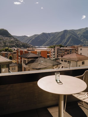 Un balcón con una mesa y un vaso de agua, con vistas a un pueblo y montañas al fondo.