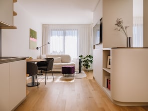A modern living room with a sofa, a dining table with chairs, a small purple ottoman, a potted plant, and a bookshelf.