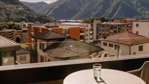 A balcony with a table and chairs, overlooking a town with buildings and mountains in the background. - THE FLAG Lugano Self Check-in (Massagno)
