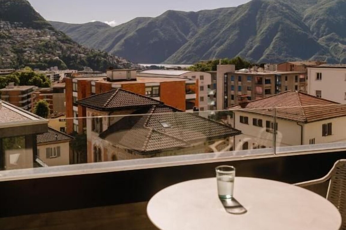A balcony with a table and chairs, overlooking a town with buildings and mountains in the background.