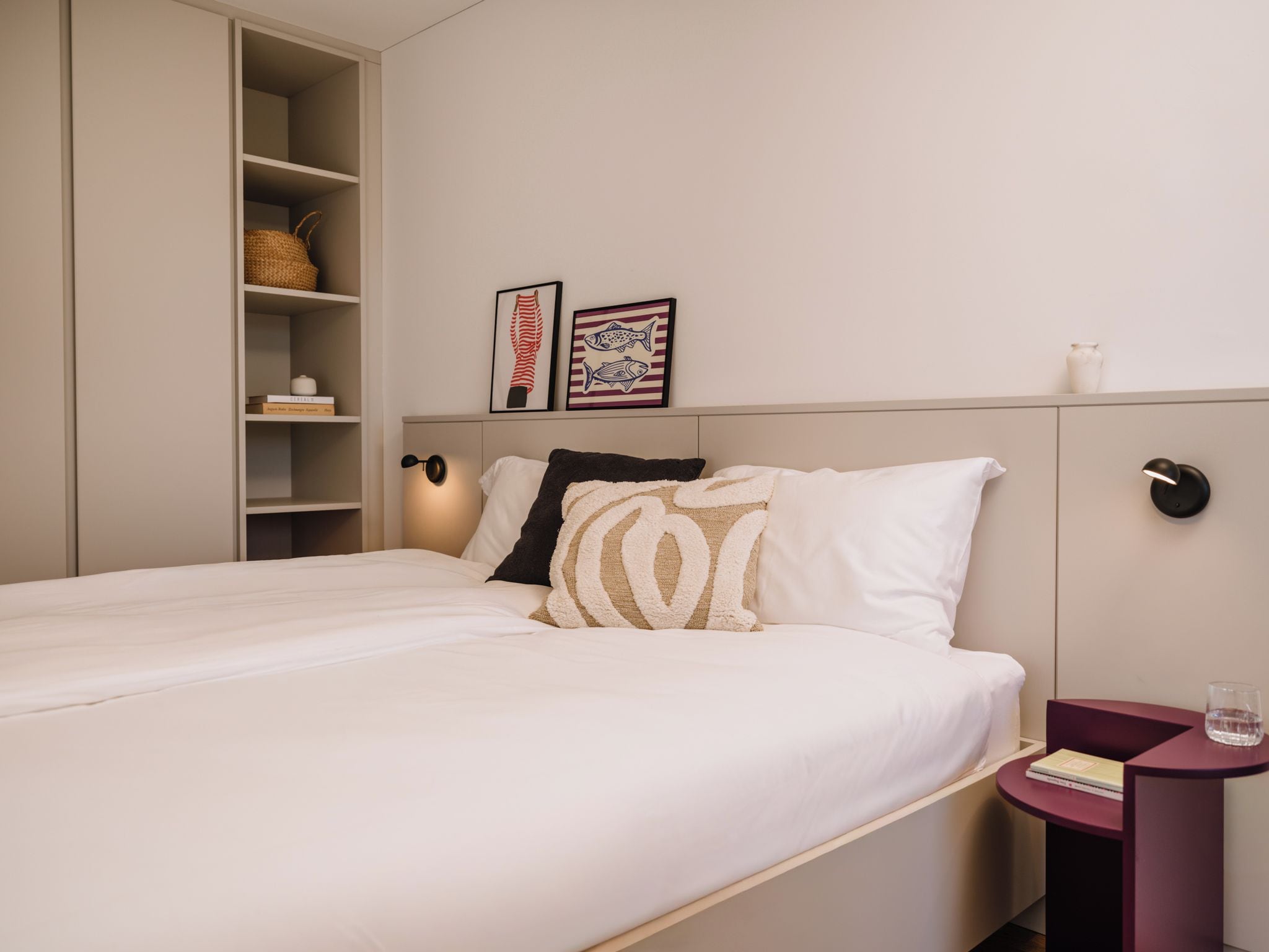 A neatly made bed with a white comforter and two pillows, a bedside table with a book and a glass, and a wall with two framed pictures.