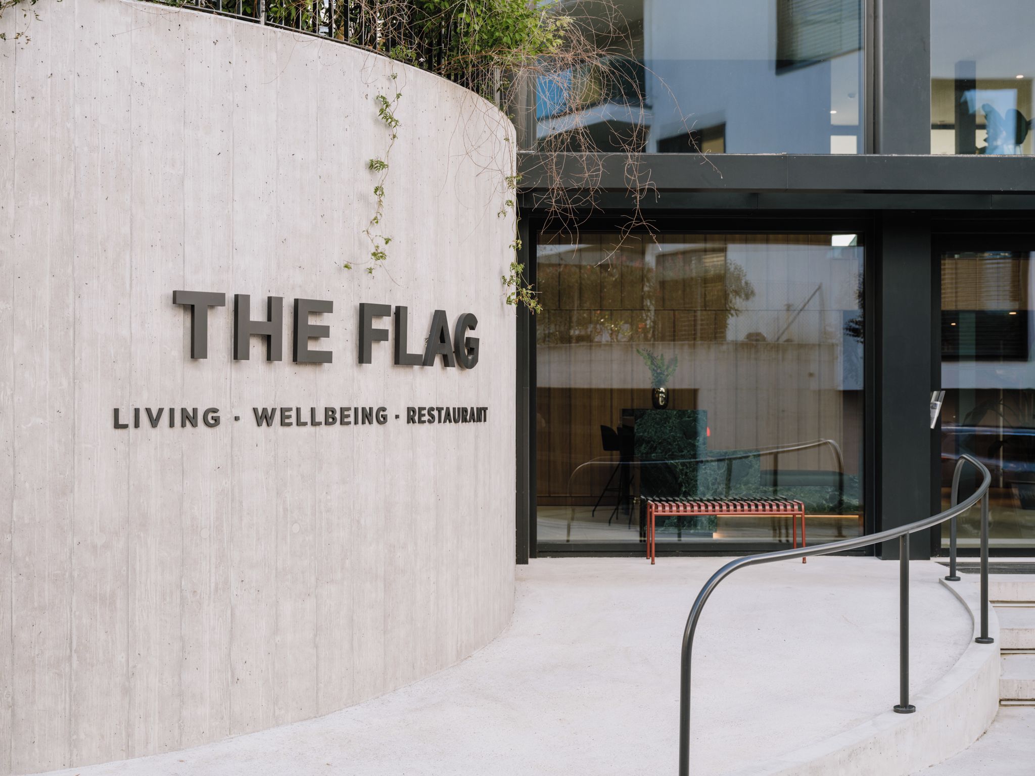 A modern building entrance with a sign reading 'THE FLAG' and a tagline 'LIVING - WELLBEING - RESTAURANT'.