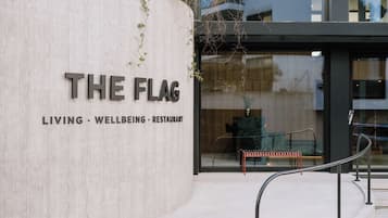 A modern building entrance with a sign reading 'THE FLAG' and a tagline 'LIVING - WELLBEING - RESTAURANT'.
