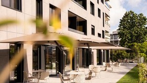Ein Außenbereich mit Tischen und Stühlen, eine überdachte Terrasse und ein Gebäude mit großen Fenstern und Balkonen.