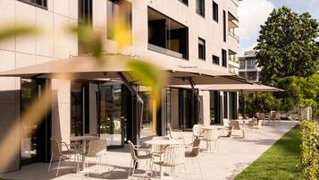 An outdoor seating area with tables and chairs, a covered patio, and a building with large windows and balconies.